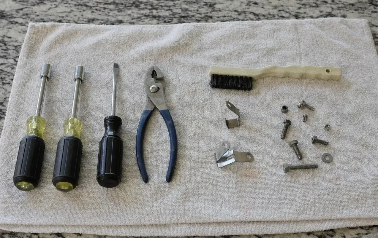Yellow-handled screwdrivers, pliers and a brush on a cloth with small clips and screws scattered beside them